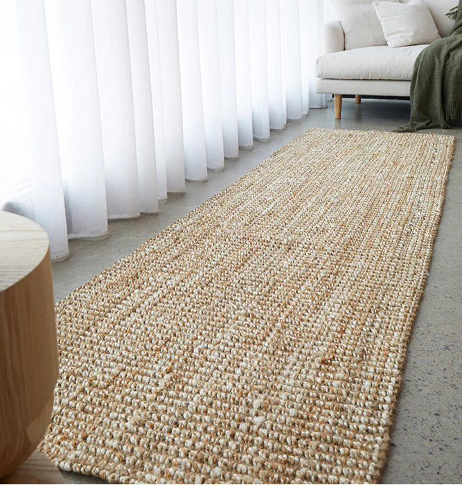 Beige woven runner rug on a light wood floor with a white sofa and green plant in the background.